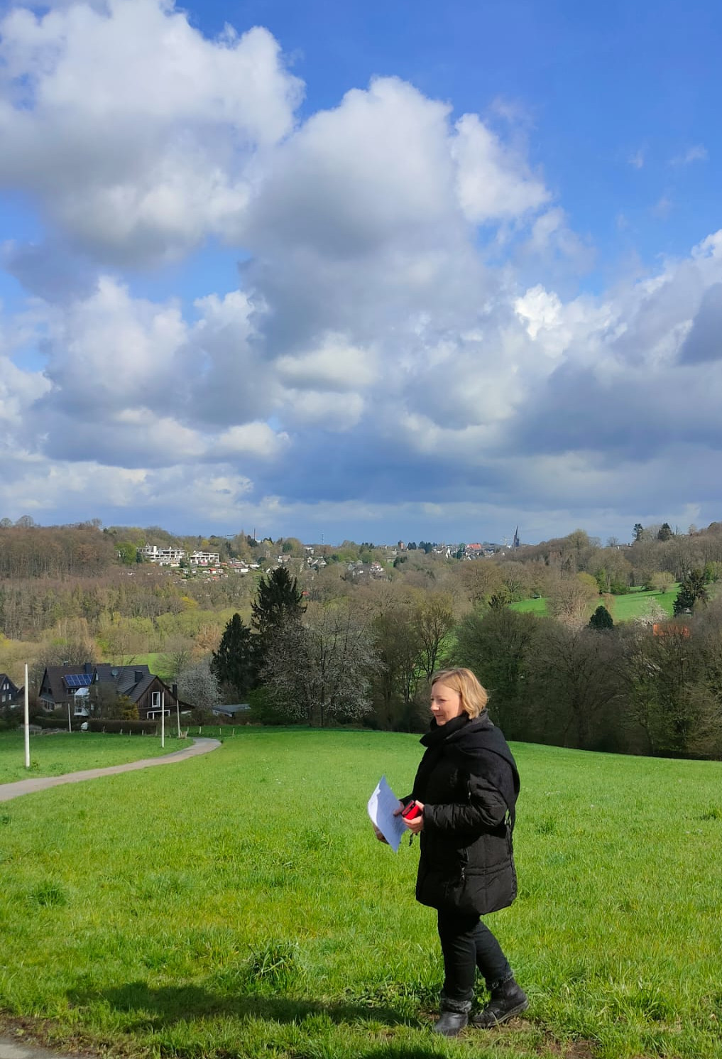 Pfarrerin Kristina Ziegenbalg auf einer saftig grünen Wiese mit Blick ins Grüntal. Der Himmel ist blau und bewölkt.
